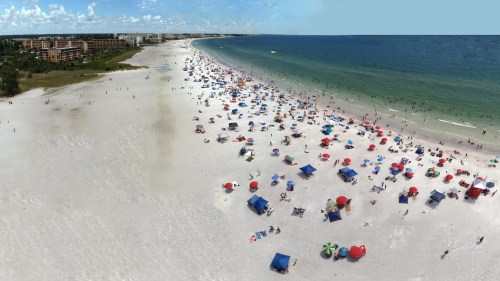 Aerial view of a busy beach with colorful umbrellas, people sunbathing, and clear blue water in the background.