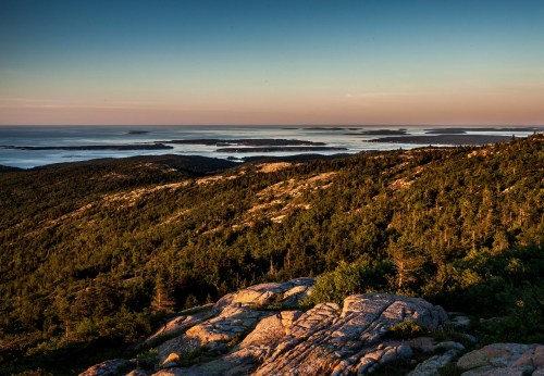 A scenic view of mountains and islands at sunset, with rocky terrain and lush greenery in the foreground.