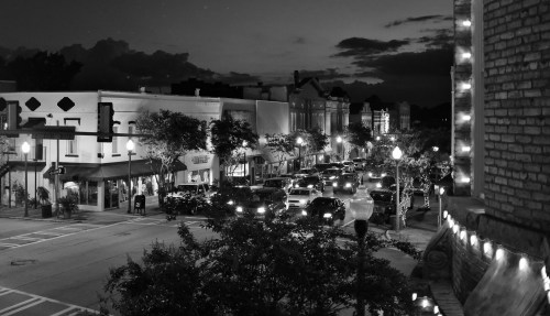 A bustling street scene at dusk, with cars and shops illuminated under a cloudy sky in black and white.