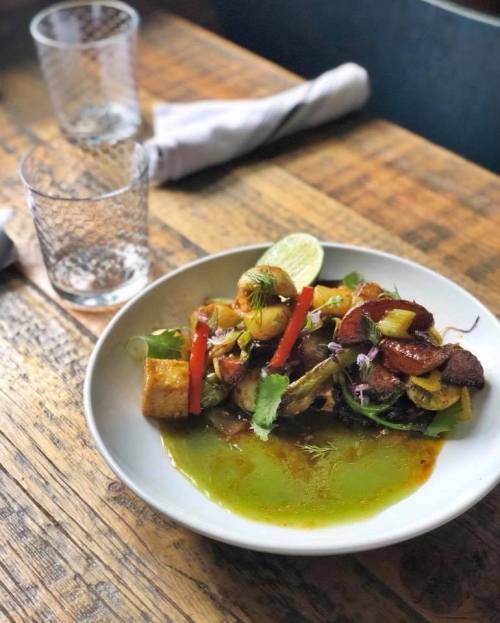 A vibrant dish featuring grilled vegetables, shrimp, and herbs on a white plate, with lime and sauce, on a wooden table.