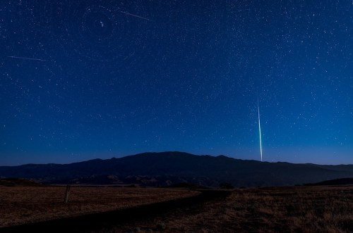 A starry night sky with shooting stars above a mountain silhouette and a winding road in the foreground.