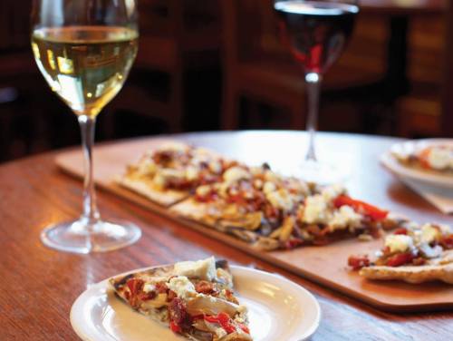 A plate of flatbread topped with vegetables and cheese, accompanied by glasses of white and red wine on a wooden table.