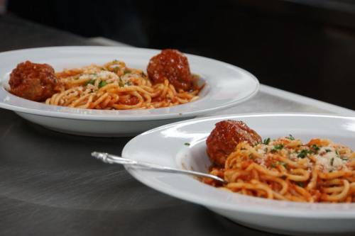 Two plates of spaghetti with meatballs, topped with grated cheese and parsley, on a stainless steel surface.
