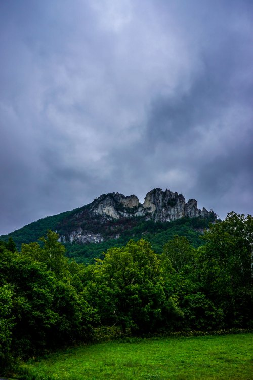 A rocky mountain peak rises above lush green trees under a cloudy sky.