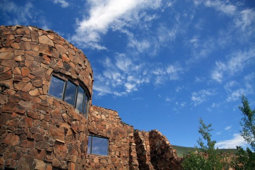 A stone building with large windows against a bright blue sky and scattered clouds. Green trees are visible nearby.