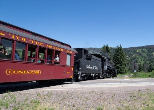 A vintage steam train passes by a red passenger car in a scenic mountainous landscape under a clear blue sky.