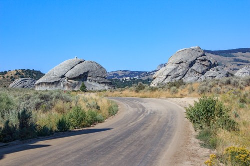 A winding dirt road leads through a landscape with large, rounded boulders and sparse vegetation under a clear blue sky.