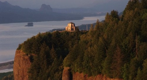 A scenic view of a cliffside building surrounded by lush trees, overlooking a river and distant mountains at sunset.