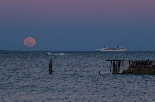 A large moon rises over the ocean, with a cruise ship and a small boat in the water, against a twilight sky.