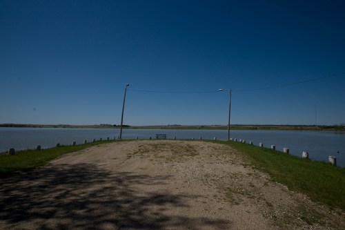 A serene lakeside view with a gravel path, green grass, and a picnic table under a clear blue sky.