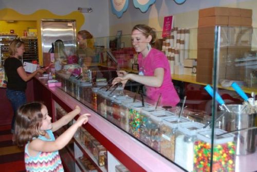 A girl points at a colorful candy display while a woman serves treats behind a glass counter in a bright shop.