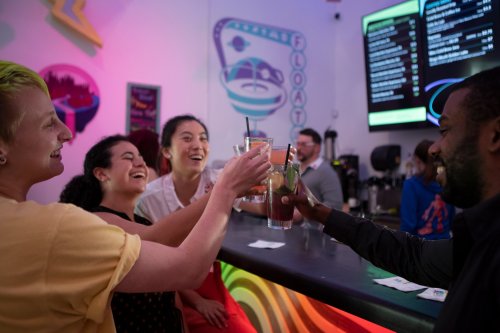A group of friends joyfully toasting with drinks at a vibrant bar, colorful lights in the background.