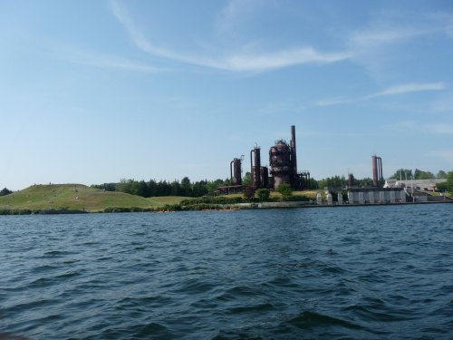 A view of an industrial facility by a lake, with green hills and people relaxing on the shore in the background.