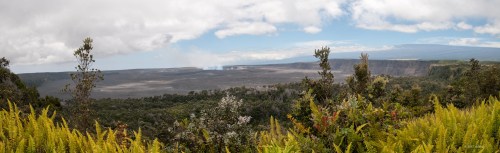 A panoramic view of a volcanic landscape with lush greenery in the foreground and a smoky caldera in the background.