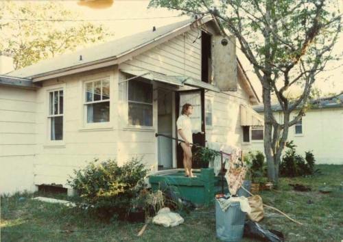 A woman stands on the porch of a modest house, surrounded by plants and a trash can, with laundry hanging nearby.