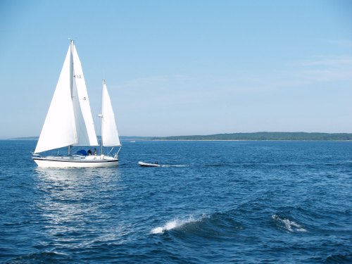 A sailboat with white sails glides across a calm blue sea, with a distant shoreline visible under a clear sky.