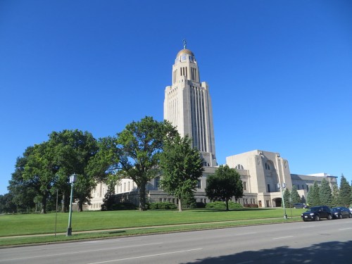 Nebraska State Capitol building with a tall tower, surrounded by green trees and a clear blue sky.