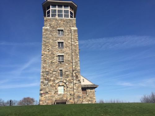 A tall stone tower with a glass observation deck against a clear blue sky, surrounded by green grass.