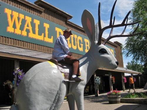 A man sits on a large, whimsical jackalope statue outside Wall Drug Store, with colorful flowers in the foreground.