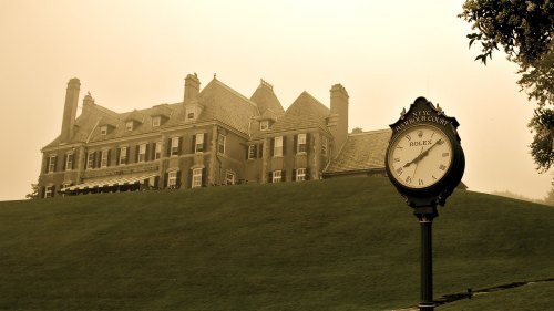 A vintage clock stands in front of a grand mansion on a grassy hill, with a foggy sky in the background.