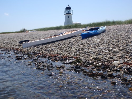 Two kayaks on a rocky shore with a lighthouse in the background under a clear blue sky.