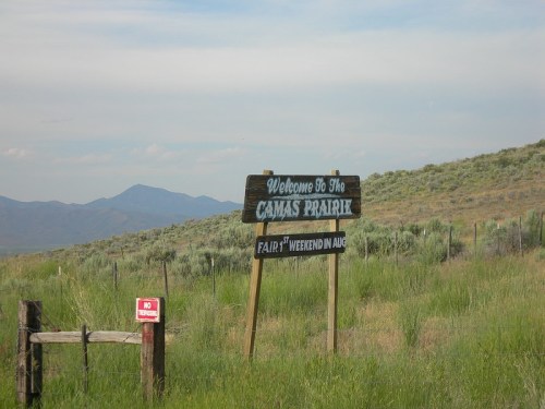 Sign welcoming visitors to Camas Prairie, with mountains in the background and green grass surrounding the area.