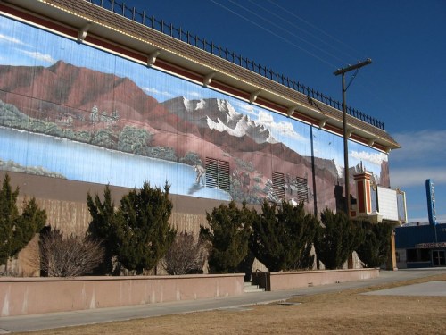Mural of mountains and a lake on a building wall, with shrubs and a clear blue sky in the background.