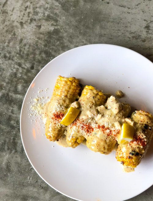 A plate of corn on the cob topped with creamy sauce, cheese, paprika, and lemon wedges on a textured surface.