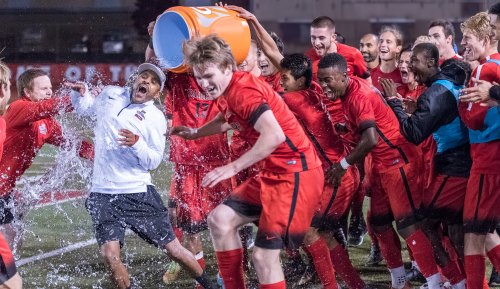 A jubilant soccer team celebrates a victory by dousing their coach with water from a cooler, surrounded by cheering teammates.
