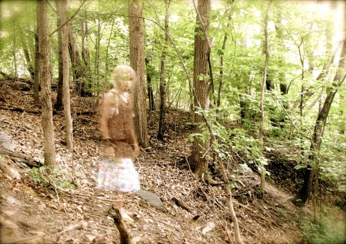 A blurred figure of a girl walking through a lush, green forest with trees and foliage surrounding her.