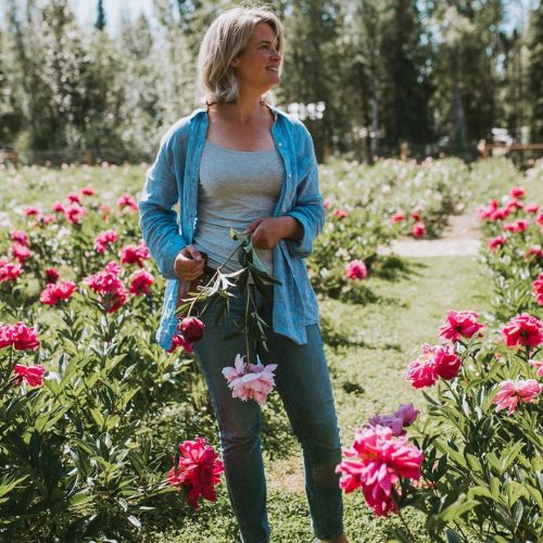 A woman stands in a flower field, holding a peony, surrounded by vibrant pink blooms and greenery.