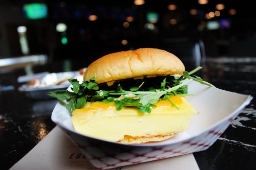 A close-up of a sandwich with a soft bun, filled with yellow cheese and fresh greens, served on a paper tray.