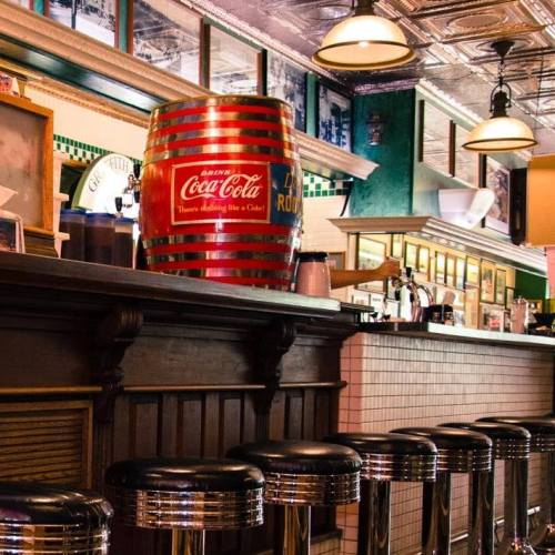 A vintage diner interior featuring a wooden bar, retro stools, and a large Coca-Cola barrel.