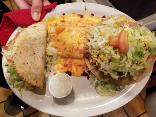 A plate with a taco, a tostada topped with lettuce and tomato, and a cheese-covered enchilada, served with onions.