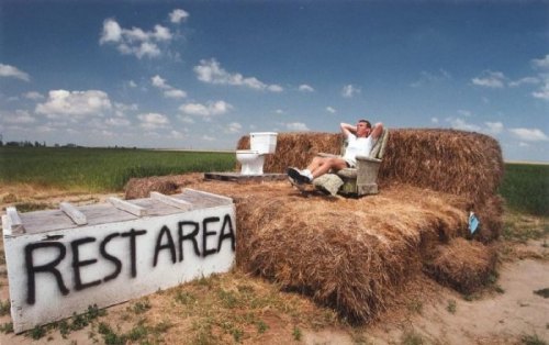 A man relaxes on a haystack couch in a field, next to a toilet, with a sign reading "Rest Area."