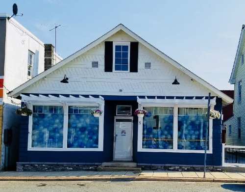 A blue and white house with large windows decorated with blue balloons, located on a sunny street.