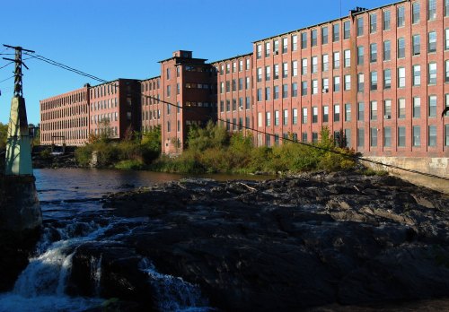 Historic red brick mill buildings along a river, with rocky banks and clear blue sky in the background.