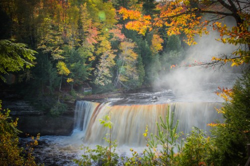 A scenic waterfall surrounded by vibrant autumn foliage and mist rising from the water.