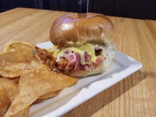 A sandwich with fried chicken, bacon, and mustard on a bun, served with crispy potato chips on a white plate.