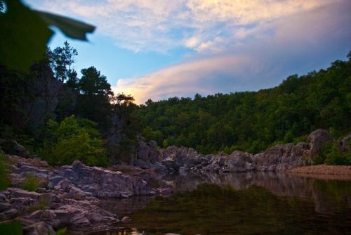 A serene river scene surrounded by rocky cliffs and lush green trees under a colorful sky at dusk.
