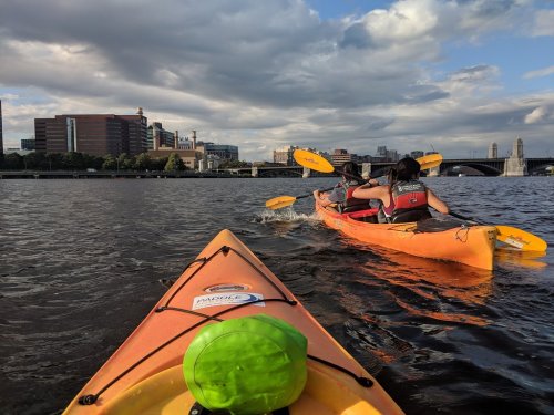Two kayakers paddle on a river with city buildings and a bridge in the background under a cloudy sky.