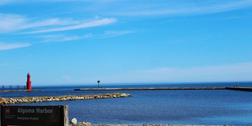 A serene view of Algoma Harbor with a red lighthouse, calm waters, and a clear blue sky.