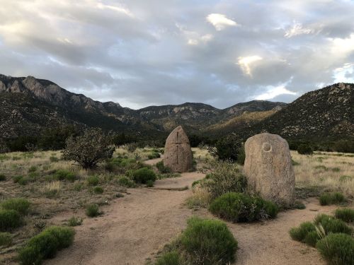 Two large stones stand on a dirt path surrounded by grass and shrubs, with mountains and cloudy skies in the background.