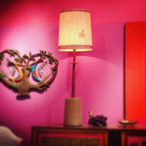 A tall lamp with a patterned shade stands against a vibrant pink wall, next to a decorative wall piece.