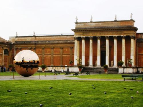 A large metallic sphere in a green courtyard, with classical architecture and columns in the background.