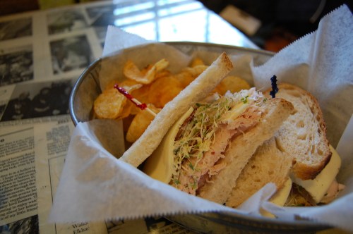 A sandwich with sprouts and turkey on bread, served with potato chips in a basket lined with parchment paper.