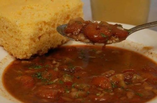 A bowl of hearty soup with vegetables and a spoonful, accompanied by a piece of cornbread and a drink.