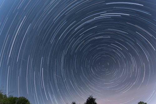 A long-exposure image of star trails forming circular patterns in the night sky, with silhouettes of trees below.