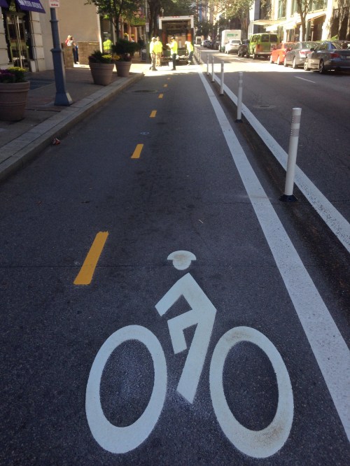 A bike lane marked with a bicycle symbol, flanked by planters and construction activity in the background.