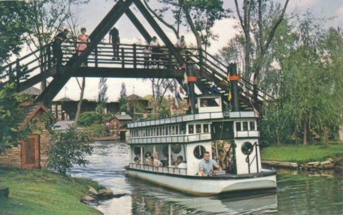 A vintage boat passes under a wooden bridge in a scenic park, surrounded by trees and greenery.